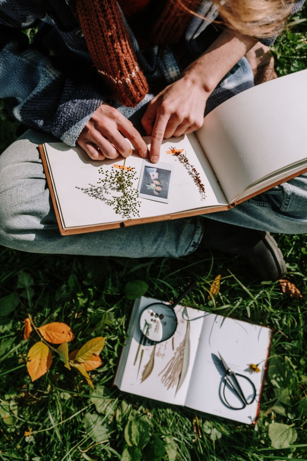 Iemand werkt buiten in het gras aan een boek met gedroogde bloemen en een schaar.